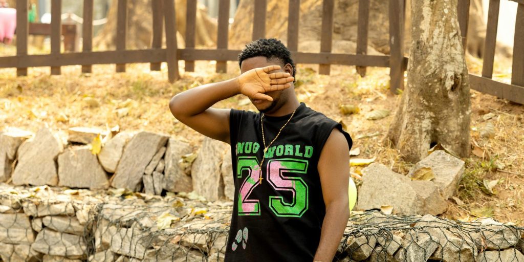 A teenager poses casually outdoors in a summer outfit, covering face with hand.
